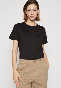 Svart bomull T-skjorte med korte ermer og rund hals, med en diskret logo trykt på brystet. Båret med beige bukser.