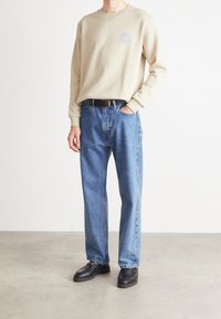 Persona che indossa una camicia a maniche lunghe beige, jeans blu dritti, cintura nera e mocassini neri, in piedi su un pavimento grigio davanti a una parete bianca.