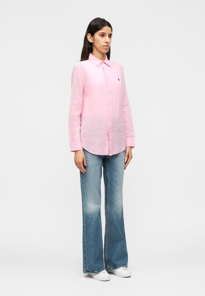 Femme debout, portant une chemise rose à manches longues boutonnée, un jean évasé bleu clair et des baskets blanches, sur un fond clair uni.