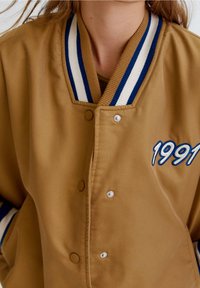 Zbliżenie osoby w brązowej kurtce varsity z niebieskim i białym paskowanym kołnierzem oraz haftem "1991" na piersi.