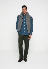 Pilkas megztinis, uždėtas ant mėlynos su sagomis marškinių, derinamas su žaliomis kelnėmis. Tamsūs batai užbaigia aprangą. Vienodas fonas.