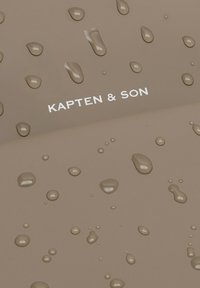 Braune strukturierte Oberfläche mit Wassertropfen und dem weißen Logo "KAPTEN & SON". Das Design zeigt verschiedene Tropfenformen und -größen.