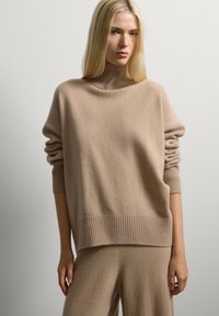 Giovane donna con lunghi capelli biondi che indossa un maglione girocollo a coste beige e pantaloni larghi coordinati su uno sfondo bianco semplice.