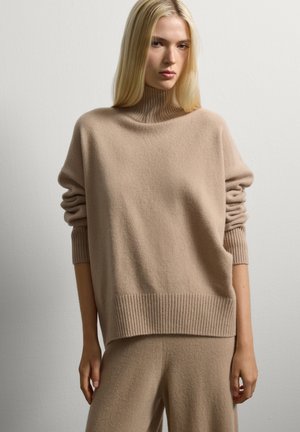 Jeune femme aux longs cheveux blonds portant un pull à col roulé beige à côtes et un pantalon large assorti, devant un fond blanc uni.