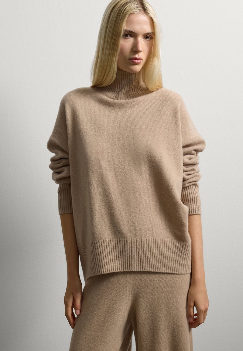 Giovane donna con lunghi capelli biondi che indossa un maglione girocollo a coste beige e pantaloni larghi coordinati su uno sfondo bianco semplice.