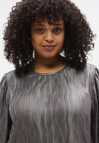 Szürke struktúrált blúz függőleges hullámos mintákkal, kerek nyakkivágással és hosszú, redőzött ujjakkal. Selymes anyag, enyhe fényű felülettel.