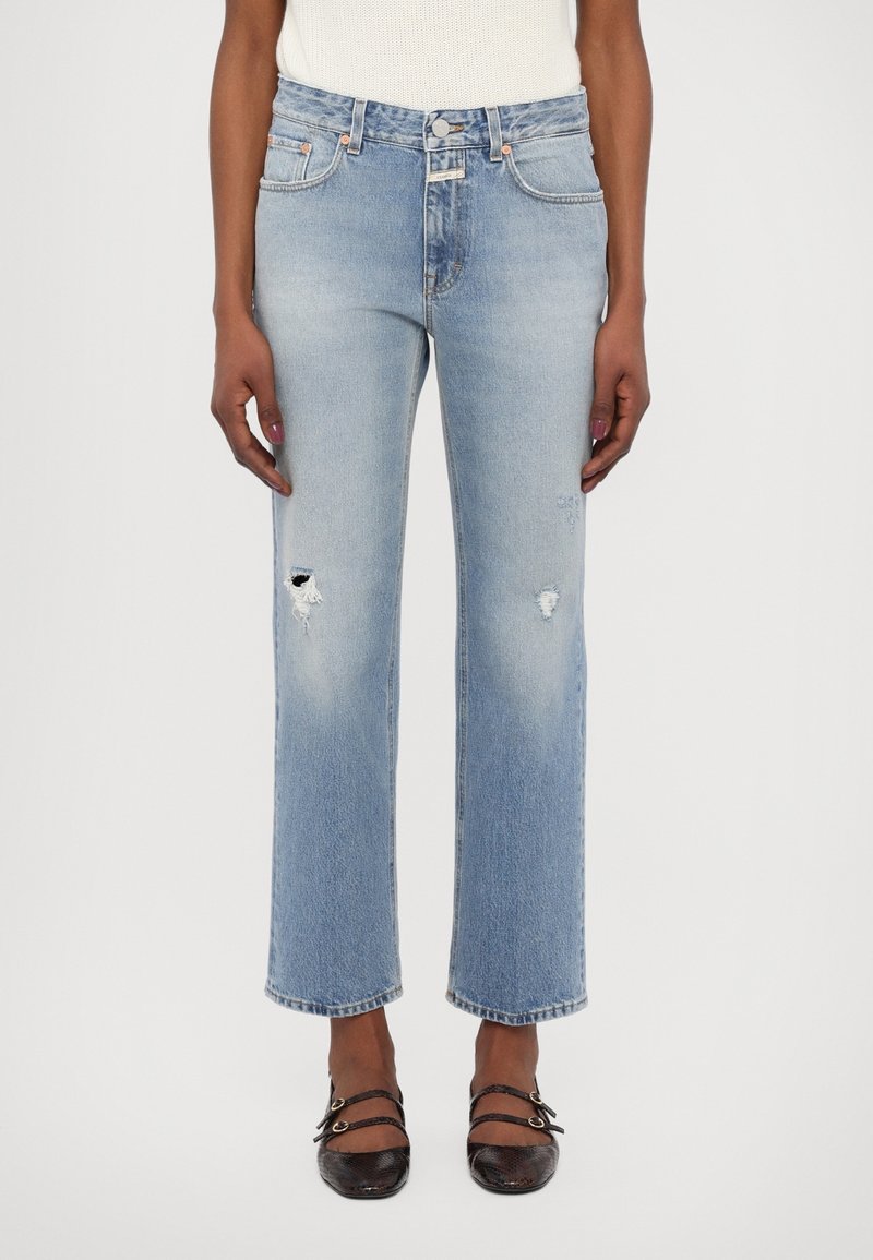 Jean droit bleu clair en denim, avec des détails usés et des ourlets effilochés, associé à des chaussures noires.