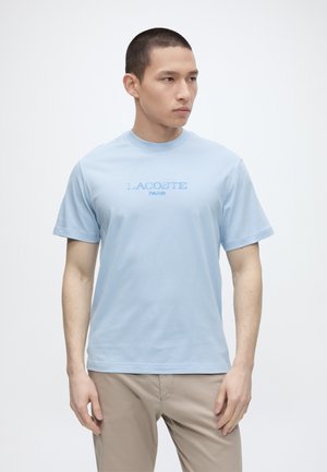 Giovane uomo che indossa una maglietta Lacoste Paris azzurro chiaro e pantaloni beige, in piedi davanti a uno sfondo bianco uniforme.