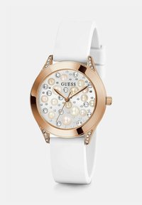 Valge rihmaga käekell roosa kullast korpusega, kristallide aksessuaaridega raami ääres ja dial, mis on kaunistatud pärlite ja kristallidega, Guess brändi logo nähtav.