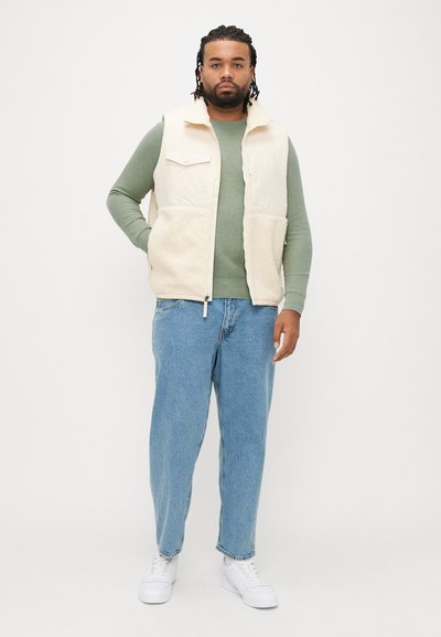 Colete de lã creme sobre uma camisola de manga comprida verde, combinado com jeans folgados azul claro e ténis brancos. Design simples e funcional.