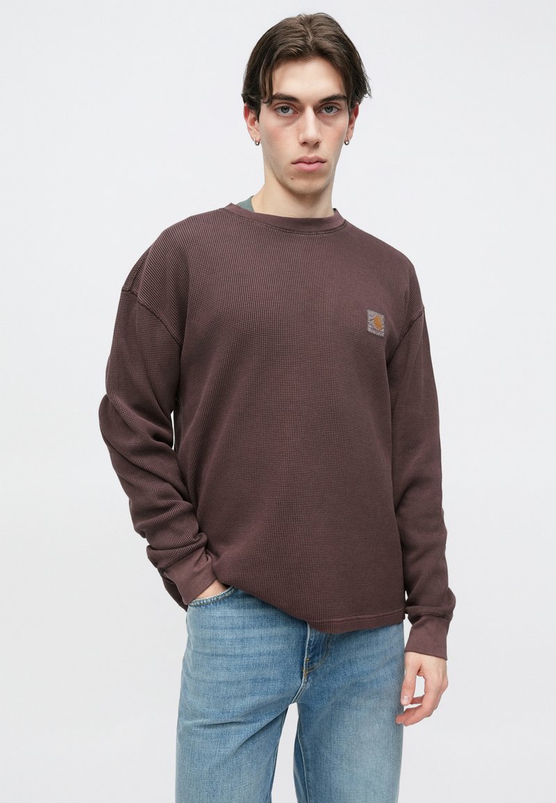 Brun ribbet langærmet skjorte med rund halsudskæring, med en lille logopatch på brystet og afslappet pasform. Båret med lyseblå jeans.