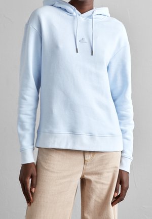 Personne portant un sweat à capuche bleu clair avec un petit logo cintre sur la poitrine et un pantalon beige sur un fond uni.