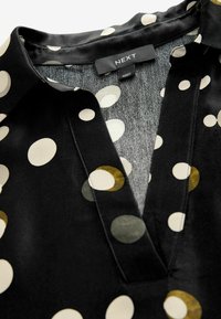 Schwarze Bluse mit großen cremefarbenen, grünen und senfgelben Kreisen. Weicher Stoff mit Kragen und einem dezenten V-Ausschnitt. Markenschild sichtbar.
