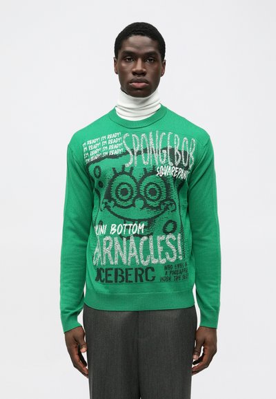 Pull en tricot vert avec un graphisme Spongebob, un texte en noir et blanc audacieux, des poignets et un col côtelés, associé à un pantalon gris.