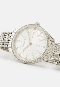 Sølv ur med en rund urskive, krystalaccenter, tekstureret hvid skive og et krystalgemt bånd med et netdesign.