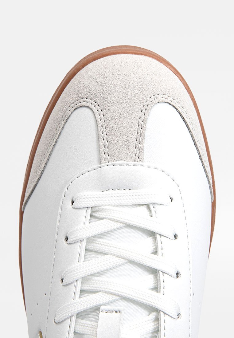 Zapatilla de cuero blanca con una puntera de ante gris, cordones blancos planos y suela de goma marrón. Diseño minimalista sin patrones visibles.