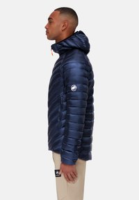 Navyblauwe pufferjack met capuchon, voorzien van een gewatteerd ontwerp, oranje ritssluiting en een logo patch op de mouw. Gladde, glanzende textuur.