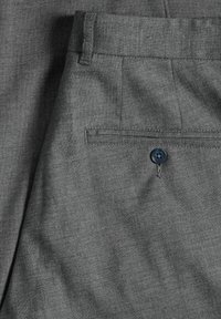 Közelkép szürke szövetből készült nadrágról, amelyen a hátsó zseb látható, rajta egy kék gomb és egy övbújtató a derékrészen.