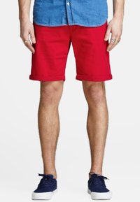 Una persona que lleva una camisa de botones azul claro y pantalones cortos rojos, de pie en zapatillas de deporte azul marino, mirando hacia la cámara contra un fondo liso.