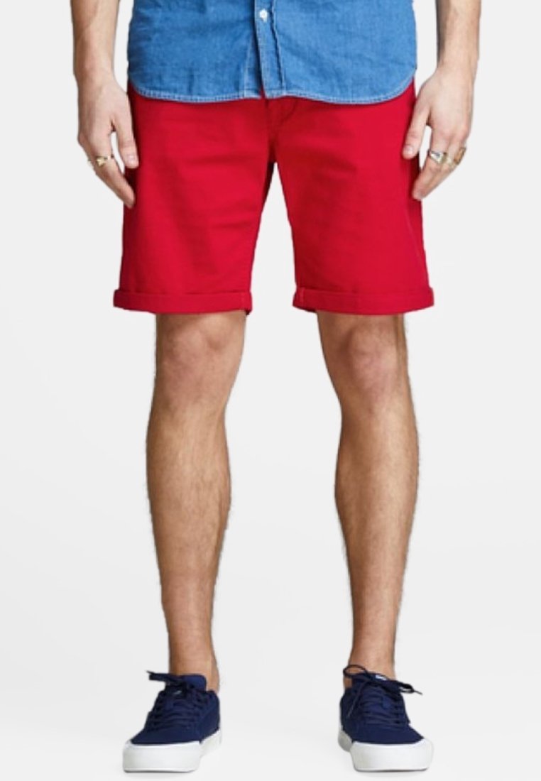 Una persona que lleva una camisa de botones azul claro y pantalones cortos rojos, de pie en zapatillas de deporte azul marino, mirando hacia la cámara contra un fondo liso.