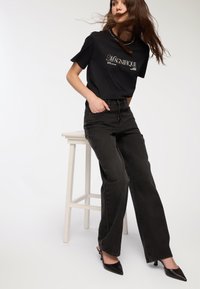 Czarny t-shirt graficzny z krótkim rękawem i napisem, w zestawieniu z czarnymi dżinsami o wysokim stanie. Czarne szpilki w szpic. Gładkie tło.