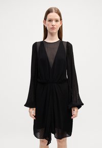 Rochie neagră cu mâneci lungi transparente, decolteu adânc în V și țesătură texturată cu buline. Talie strânsă cu un detaliu drapat în față.