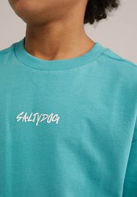Turkusowy bawełniany sweatshirt z okrągłym dekoltem i ściągaczami, z napisem "SALTYDOG" na białym tle z przodu. Gładka tekstura.