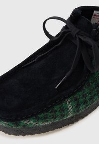 Scarpa alla caviglia in suede nero con bordi in lana a motivo verde e grigio, suola in gomma testurizzata e lacci neri fissati con aghetti metallici.