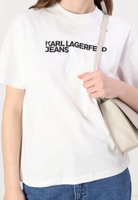 Vit bomulls-t-shirt med korta ärmar med svart text "KARL LAGERFELD JEANS." Kompletteras med en ljusgrå handväska.