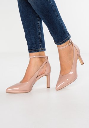 Szpilki z lakierowanej skóry w kolorze nude, z ostrym noskiem, paskiem wokół kostki i zwężającym się obcasem typu szpilka, zestawione z niebieskimi dżinsami.