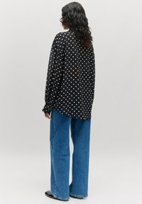 Zwarte blouse met witte stippen, lange mouwen, doorschijnende stof, ontspannen pasvorm, gecombineerd met hoge taille blauwe wijde jeans en zwarte schoenen.