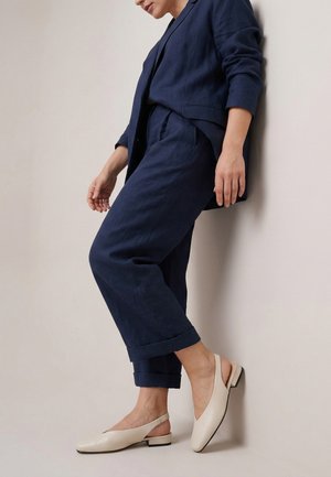 Kobieta w granatowym garniturze i beżowych butach na niskim obcasie typu slingback, opierająca się o jasną ścianę z jedną nogą zgiętą.