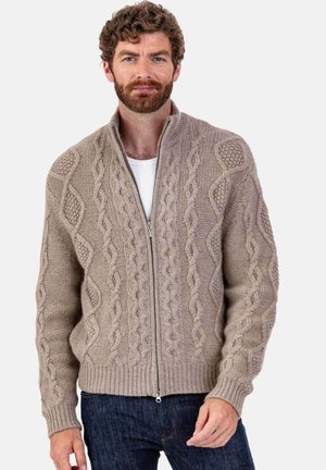 Beige knitted cardigan with a zip front, featuring cable and ribbed patterns, chunky texture, and a relaxed fit. Styled over a white shirt.