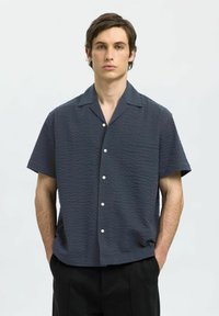 Kortermet, mørkeblå skjorte med strukturt mønster, knappet front og camp krage, sammen med svarte bukser.