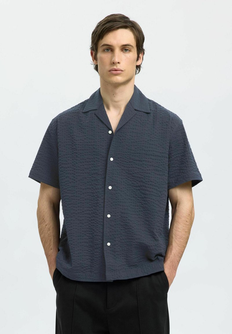 Kortermet, mørkeblå skjorte med strukturt mønster, knappet front og camp krage, sammen med svarte bukser.