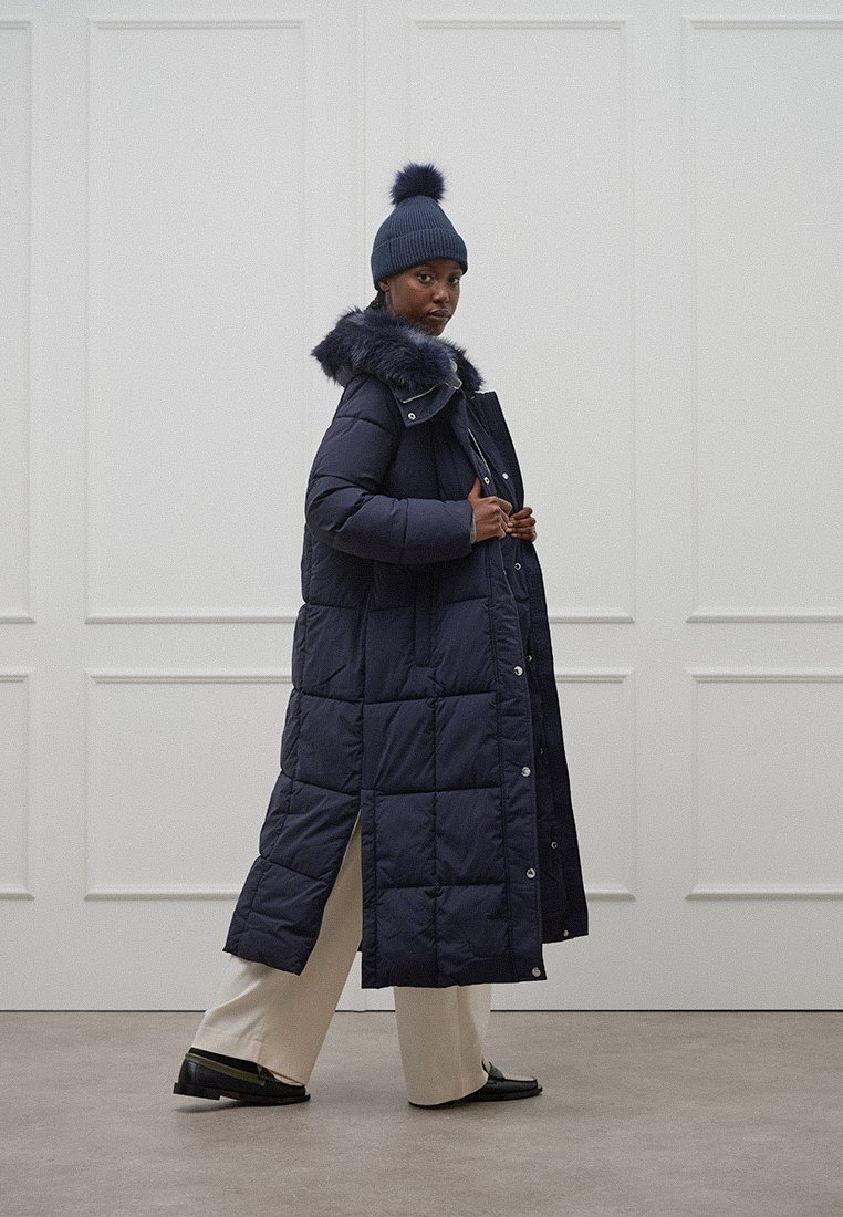 Donna che cammina all'interno indossando un lungo cappotto imbottito blu navy con collo in pelliccia, un berretto blu navy con pompon, pantaloni larghi crema e scarpe nere.