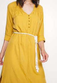 Żółta sukienka z guzikami, z dekoltem w kształcie litery V, rękawami o długości do łokci i białym sznurkiem w talii. Posiada teksturowany gorset i zwiewny materiał.
