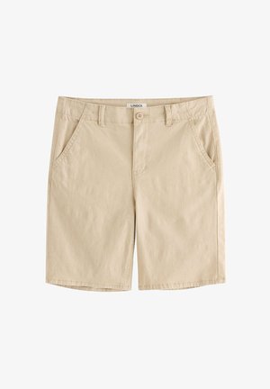 Dreng med rødt hår står med hænderne i lommerne, iført navyblå kortærmet skjorte, beige shorts, hvide sokker og hvide sneakers på hvid baggrund.