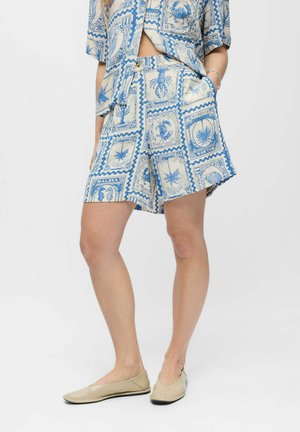 Osoba nosiaca modré a béžové šortky a košeľu s vintage motívmi známok so morskými tvormi, v kombinácii s béžovými balerínami na bielom pozadí.