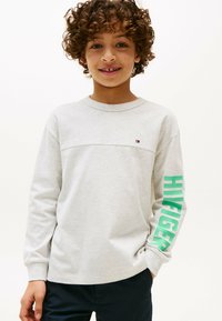 Lys grå langærmet T-shirt med et lille logo på brystet og fed grøn tekst "HILFIGER" på venstre ærme. Blødt stof.