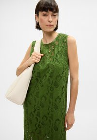 Γυναίκα με κοντά καστανά μαλλιά, φορώντας ένα αμάνικο πράσινο δαντελένιο φόρεμα, κρατώντας μια μεγάλη κρεμ τσάντα ώμου απέναντι από έναν απλό φόντο.