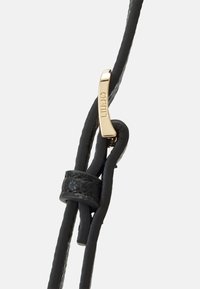 Schwarzes Lederarmband mit glatter Textur, ausgestattet mit einem goldfarbenen Metallelement, das mit "LIU JO" graviert ist, und einem geknoteten Abschnitt zur Verstellbarkeit.