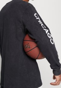 Svart långärmad skjorta med "CHICAGO" tryckt i vitt på vänster ärm. Bäraren håller en basketboll mot sin kropp.