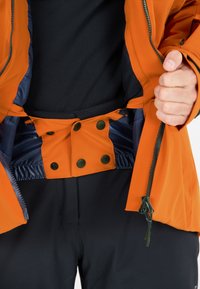 Orange vandtæt jakke med lynlås, trykknapper og elastisk indvendig. Fastgjort til sorte bukser. Glat tekstur.