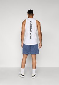 Balta be rankovių marškinėlių, ant nugaros juodu vertikaliai užrašytas "UNDER ARMOUR", derinami su mėlynomis sportinėmis šortais ir sportiniais bateliais.