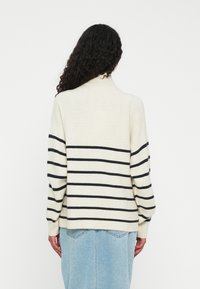 Personne avec de longs cheveux bouclés portant un pull crème à col roulé avec des rayures noires horizontales et une jupe en denim bleu clair, vue de dos.