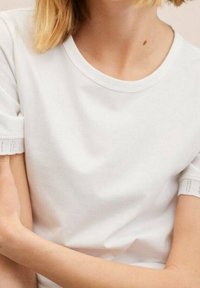 Camiseta blanca de manga corta con una textura suave, cuello redondo y detalle de encaje en los dobladillos de las mangas. Tejido ligero con un corte informal.