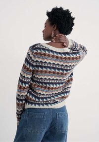 Moteris su trumpais garbanotais plaukais, vilkinti raštuotą megztinį ir mėlynus džinsus, kairiąja ranka liestinėja savo kaklą, atsisukusi į šoną.