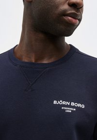 Κοντινό πλάνο ενός ατόμου που φοράει μπλε σκούρο φούτερ με το κείμενο «BJÖRN BORG STOCKHOLM 1956» τυπωμένο σε λευκό στο στήθος.