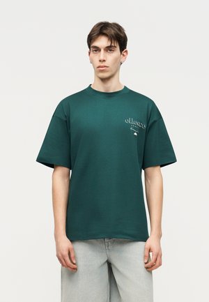 Νέος άνδρας με κοντά σκούρα μαλλιά που φοράει ένα μεγάλο μπλε-πράσινο T-shirt με μικρό λευκό λογότυπο στο στήθος και ανοιχτό γκρι τζιν, όρθιος.